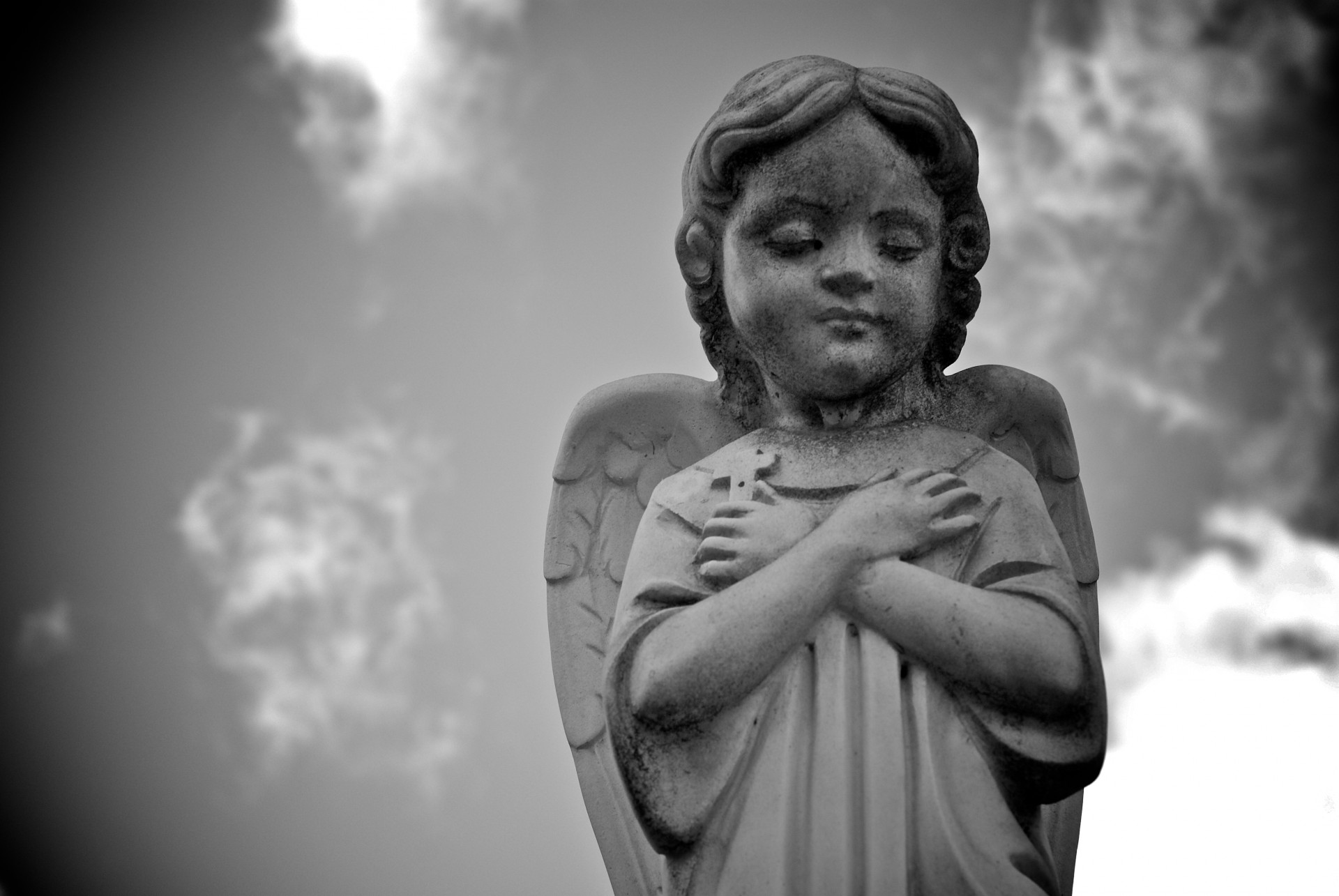 Statue of a child angel stands beneath the sky with its arms crossed across its chest.