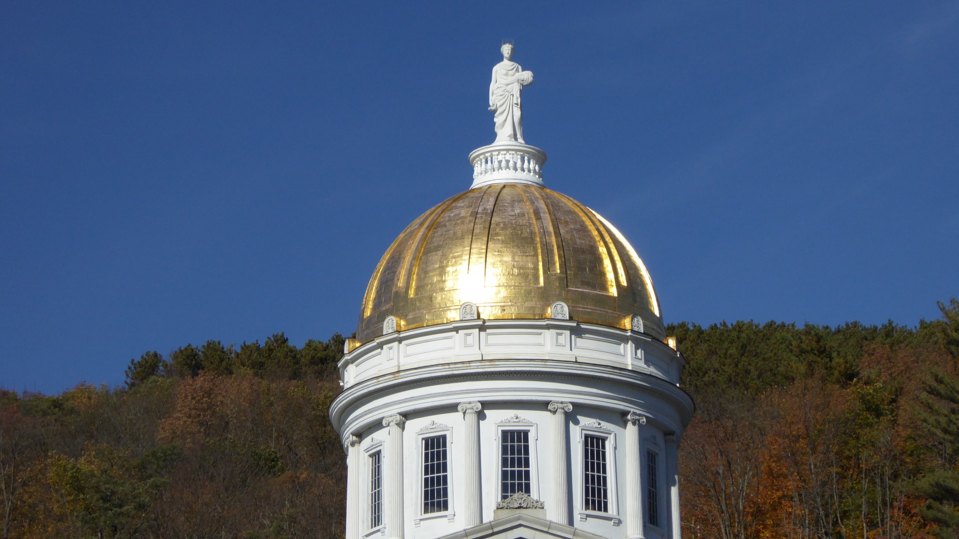 Capital building in Montpelier Vermont with a gold dome and Greek statue. Autumn foliage in the background