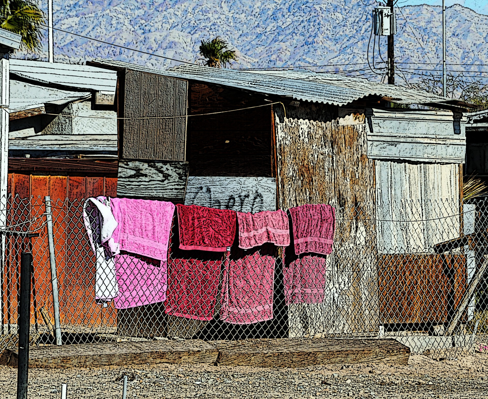 4 towels in different shakes of pink hang to dry on a chain link fence that surrounds a home that is nearly in ruins.  Taken at Bombay Beach, California. Artistic effect applied