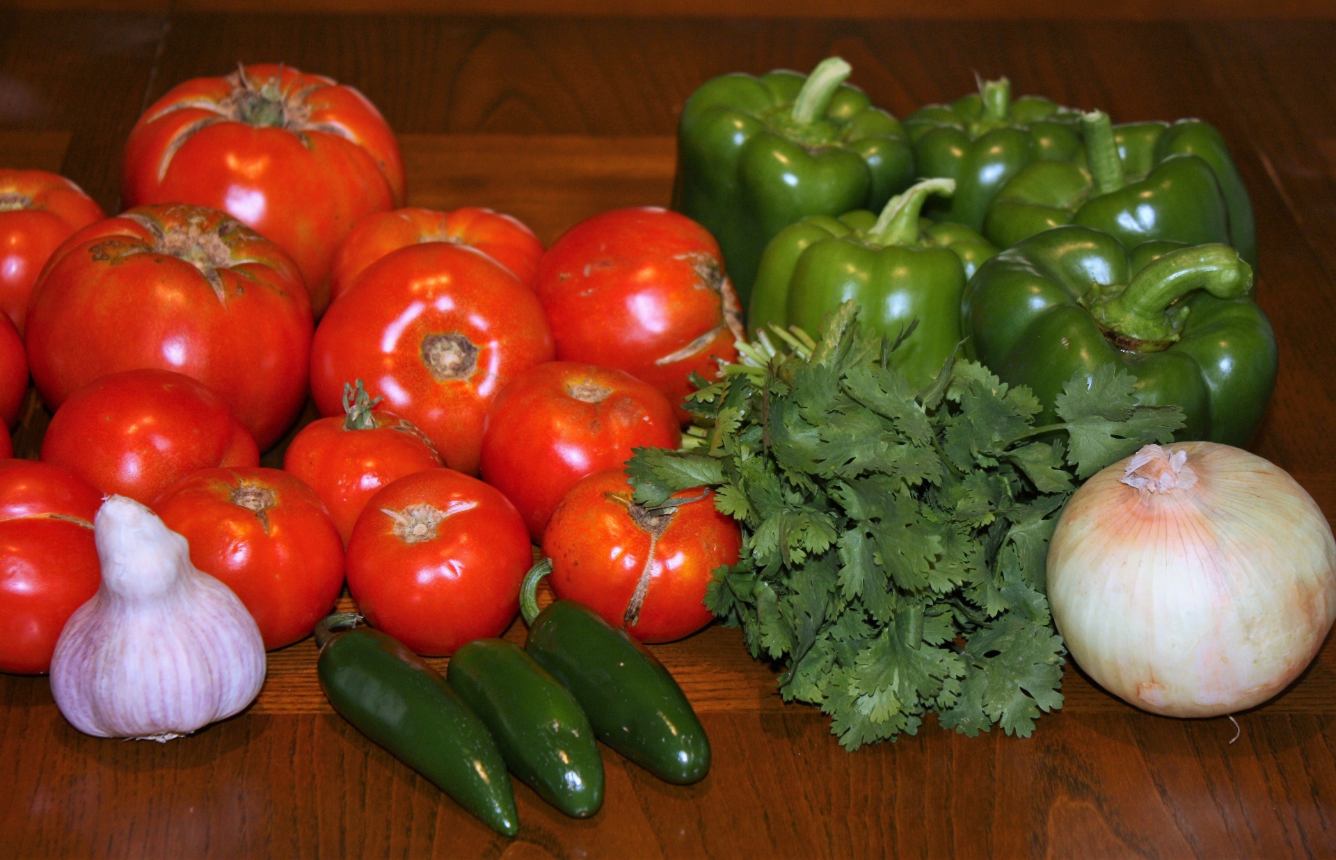 Fresh vegetables, tomatoes, cilantro, jalapeno peppers, onion, garlic, and bell peppers arranged on a brown kitchen table. These are ingredients for making homemade picante sauce.