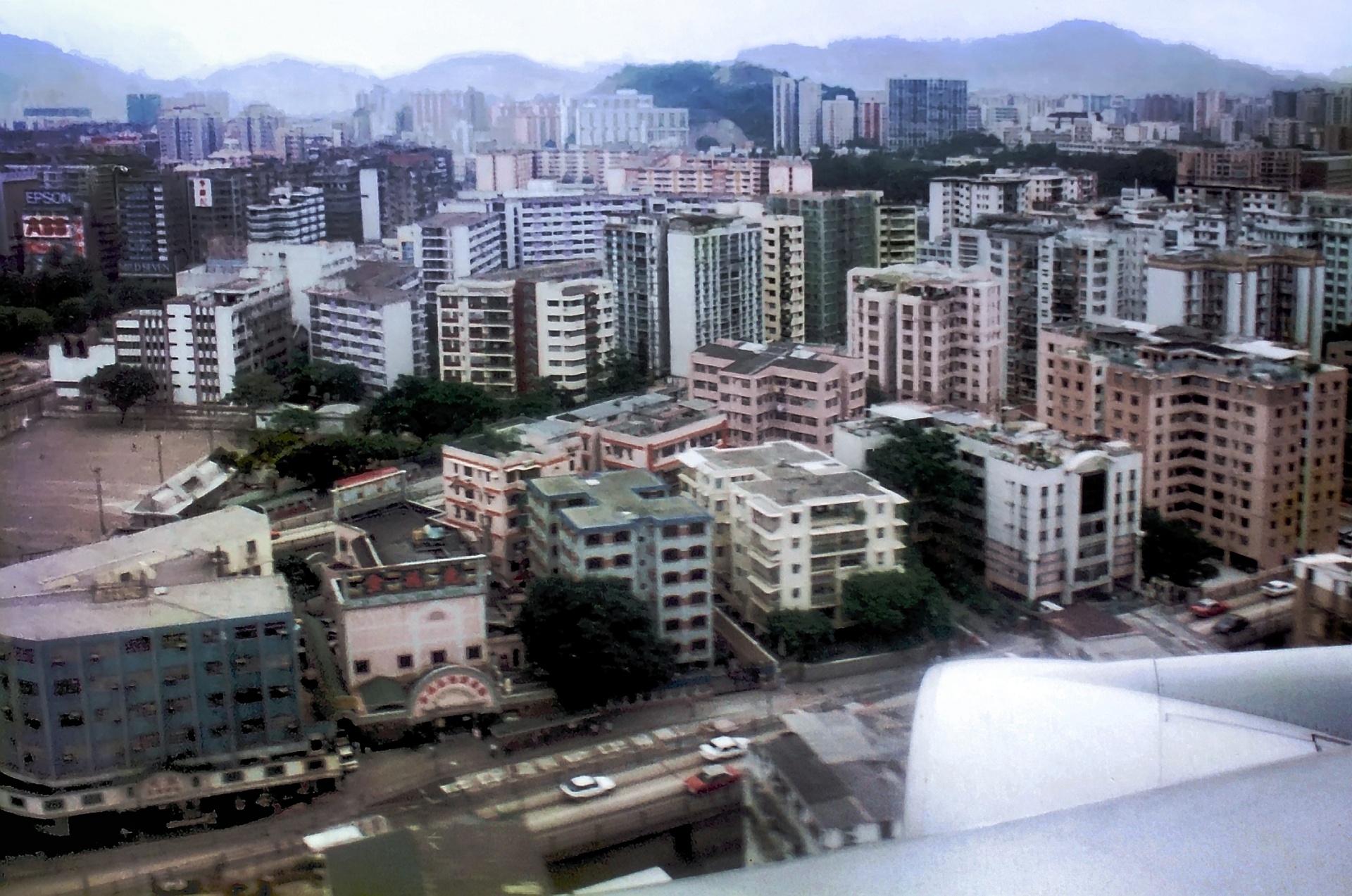 Plane coming in to land at the former Kai Tak International Airport. At less than 150 metres, shop signs are clearly visible. In front of the pink building towards the bottom left of the picture a bus stop can even be seen, with people waiting at it.