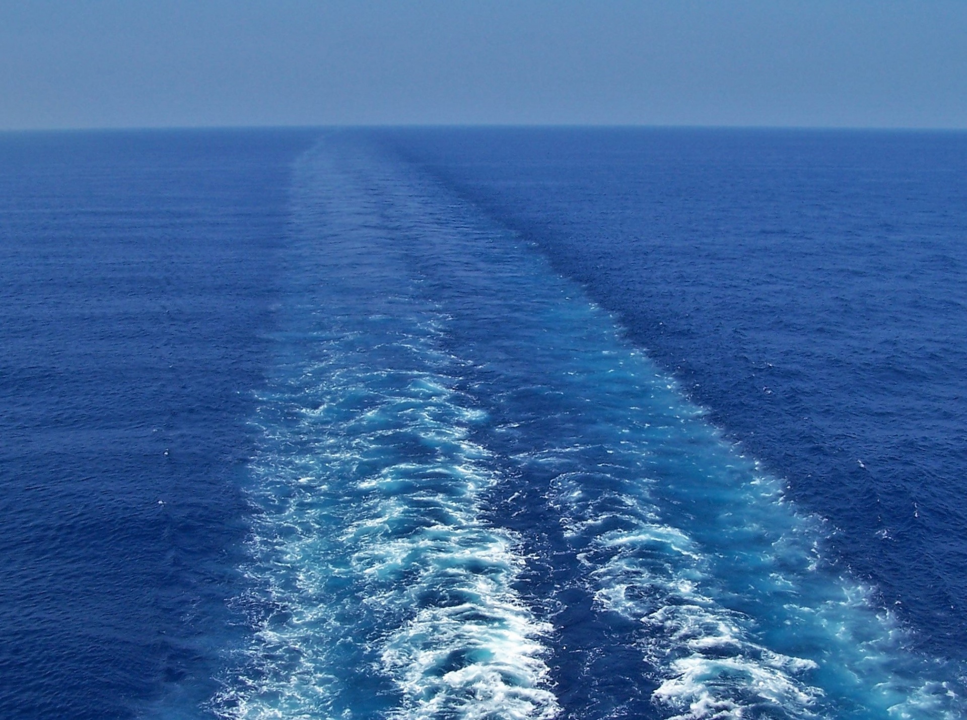 The wake from behind a cruise ship in the beautiful blue water of the Pacific Ocean as it trails to meet a beautiful clear blue sky.