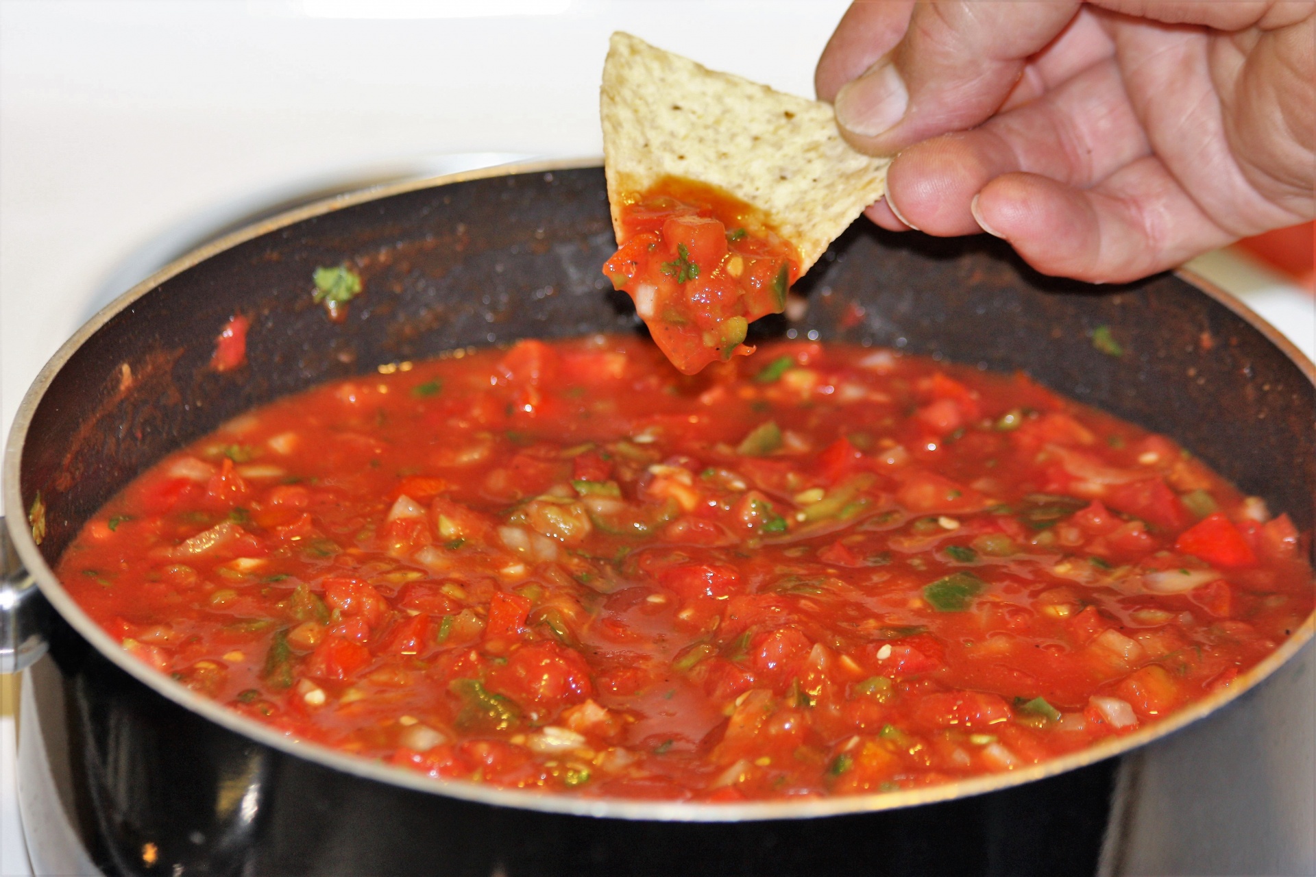 A man's hand is holding a tortilla chip that he has dipped into a pot of home-made picante sauce that is cooking on a stove.