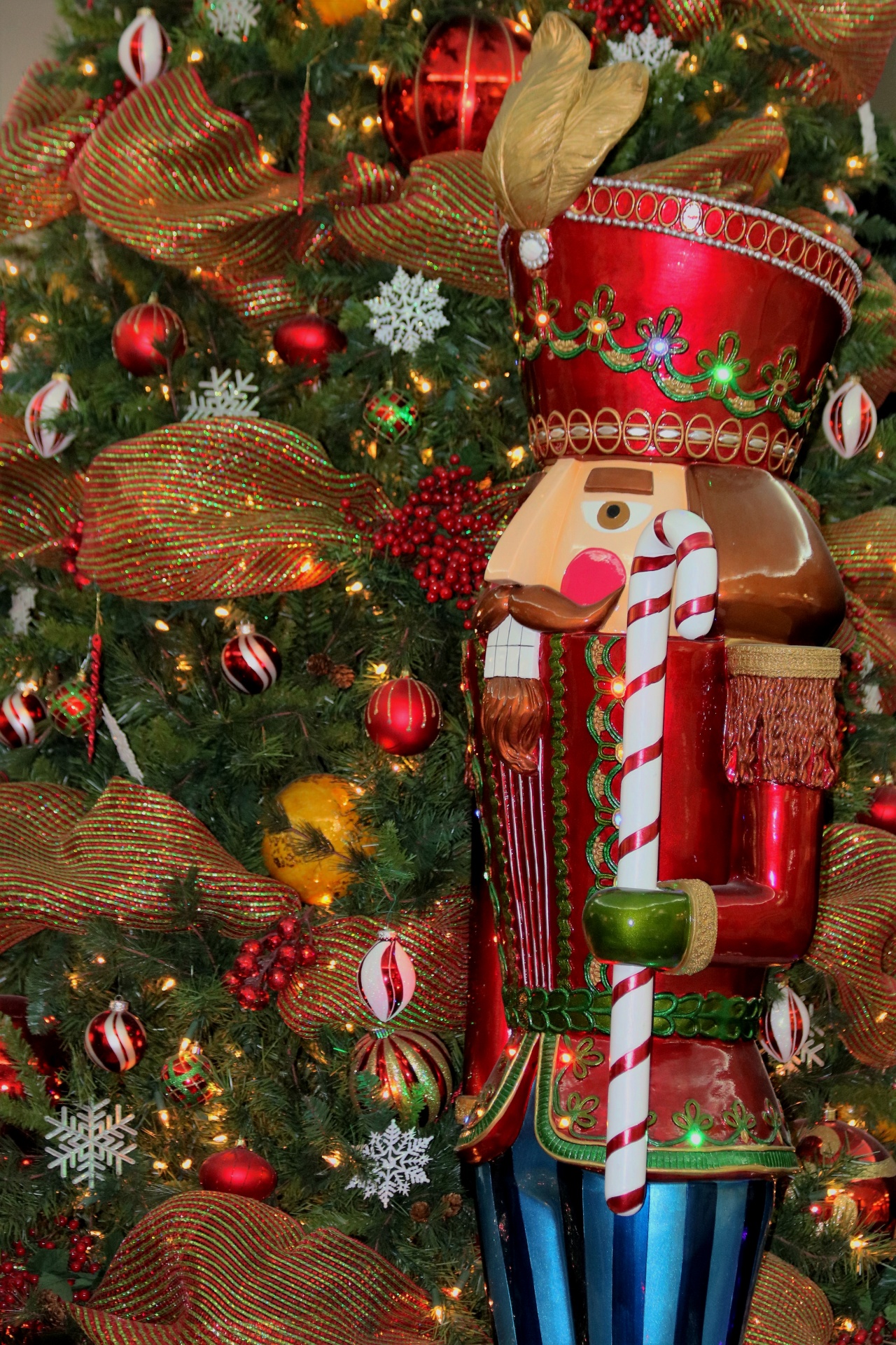 A large nutcracker soldier stands beside a green Christmas tree that is decorated with red and gold ribbon, red berries and red and white Christmas balls.