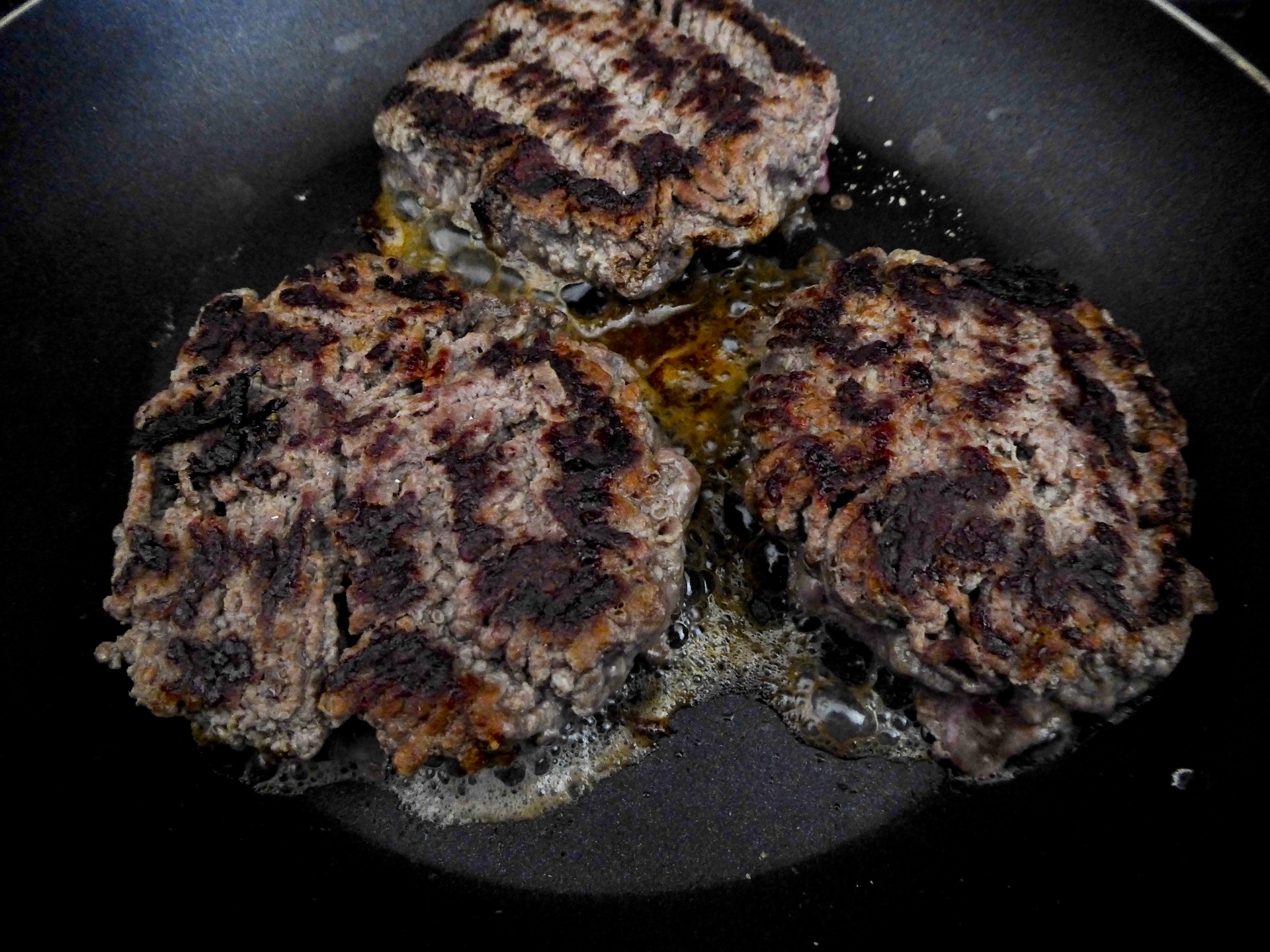 Three hamburgers sizzling as they cook in a frying pan indoors