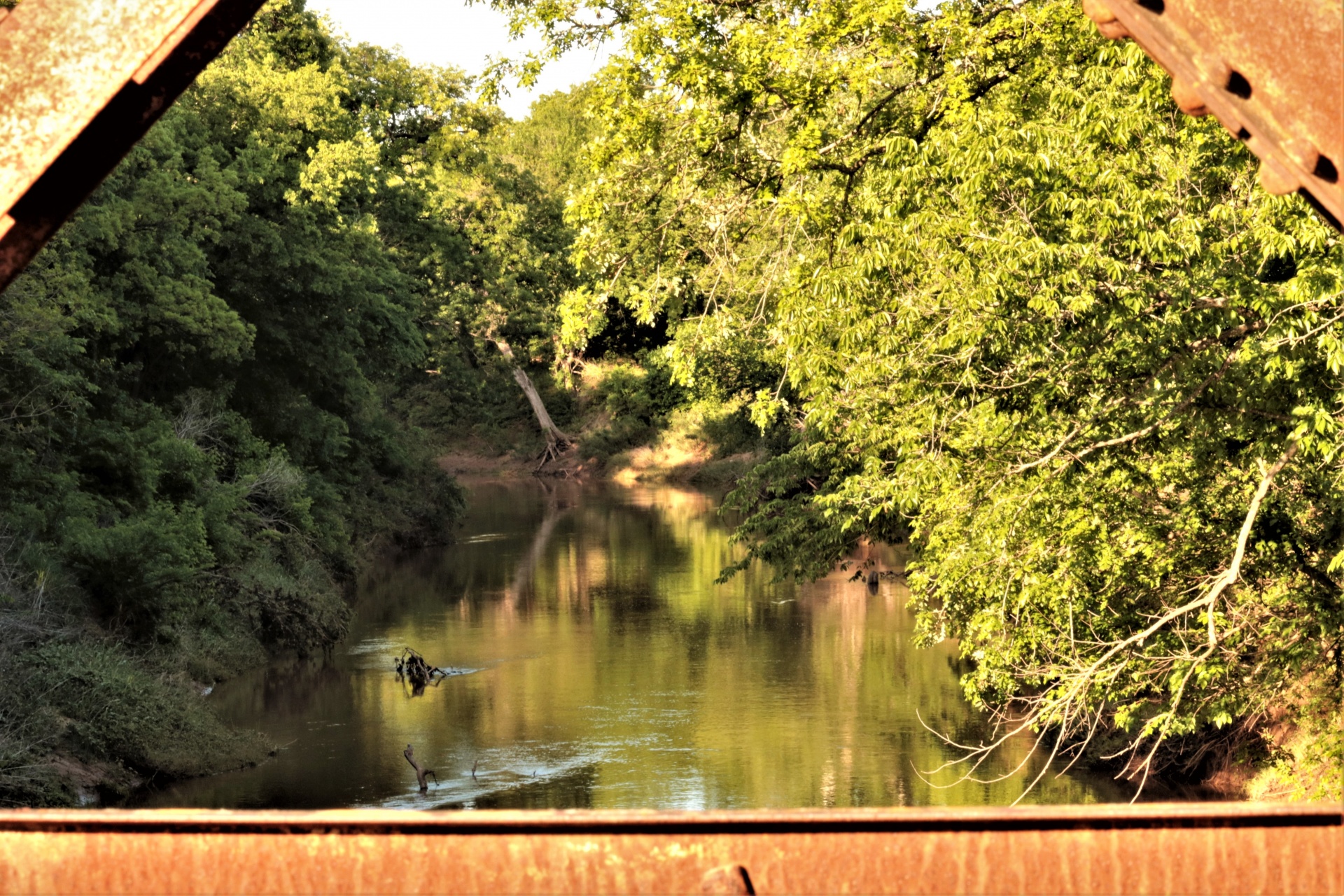 The view of a shimmering creek as it flows through green summer trees, as seen through the trusses of an old rusty bridge, in summer.