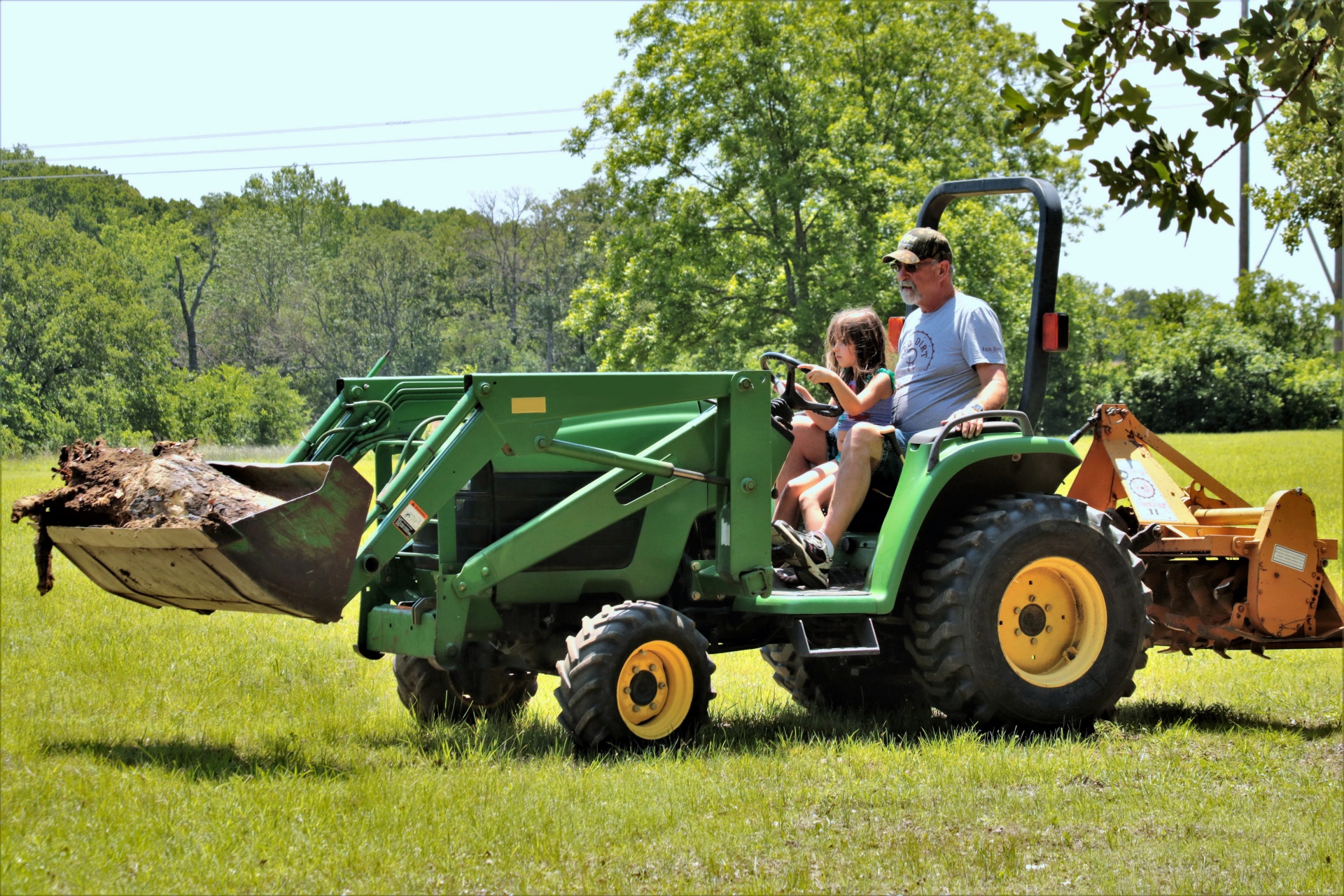 A cute little girl, sitting in her grandfather's lap, driving a farm tractor in a green country field.