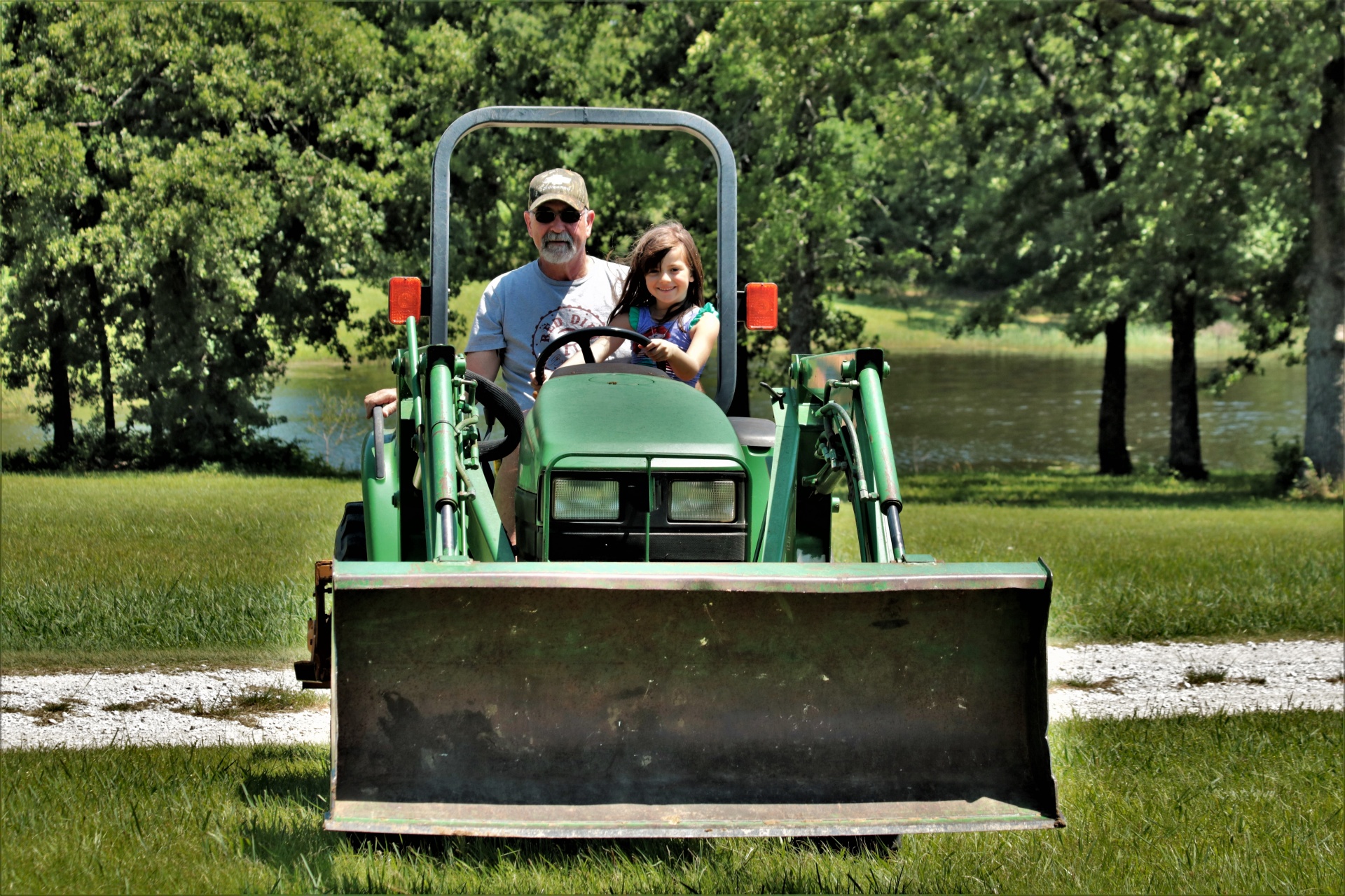 A cute little girl and her grandfather driving a farm tractor in a green grassy country field with trees and a farm pond in the background.
