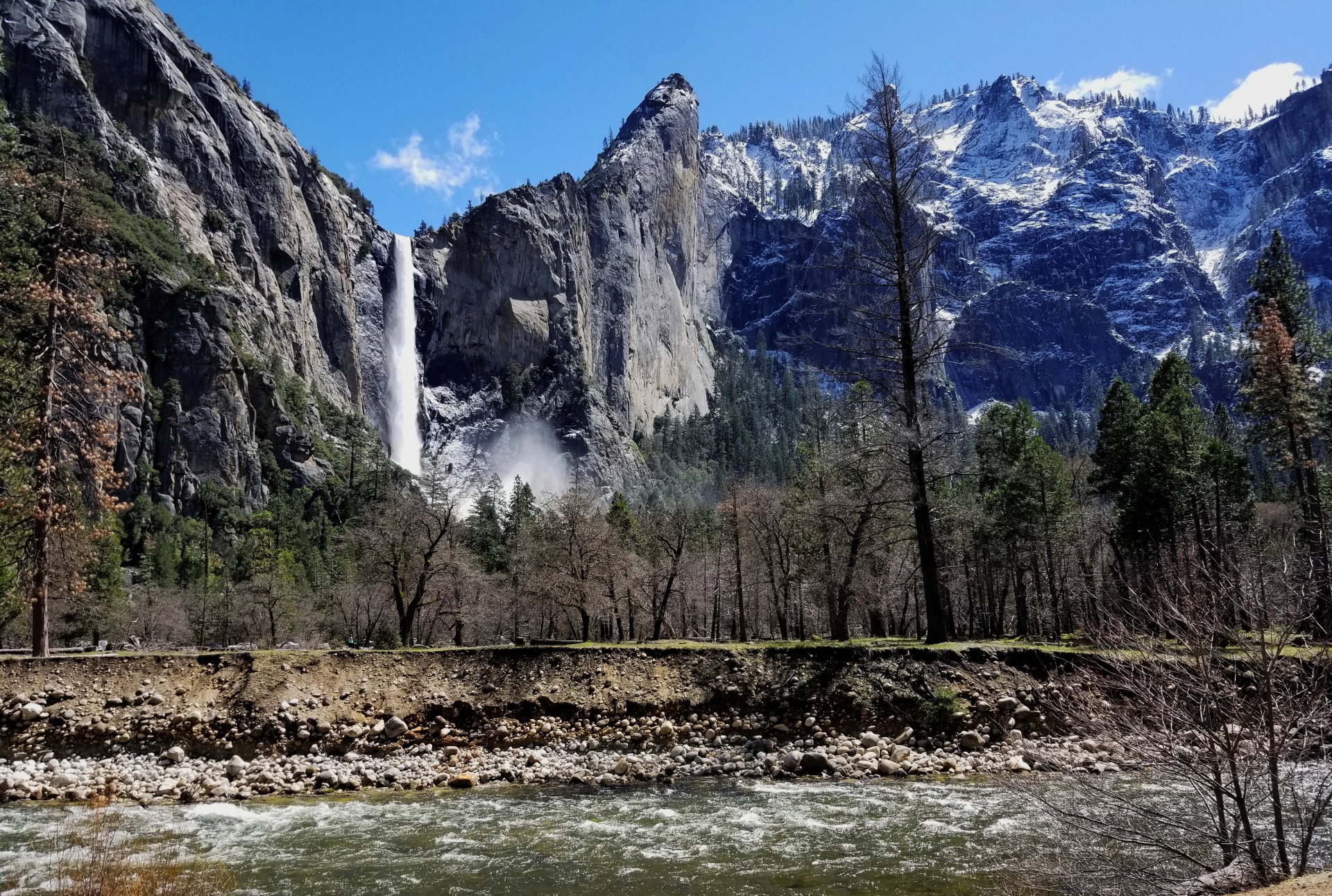 Merced River flows next to the road with waterfalls falling from snow capped mountains in Yosemite.