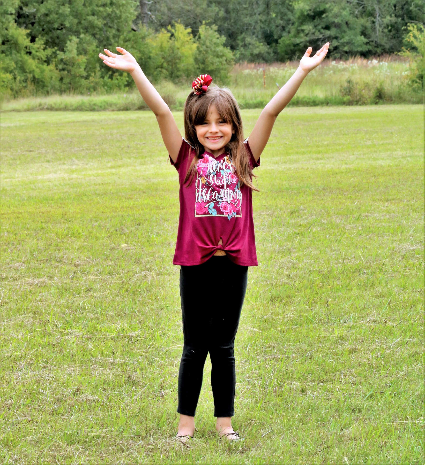 A cute smiling little girl is standing in a green country field with her arms spread out over her head.