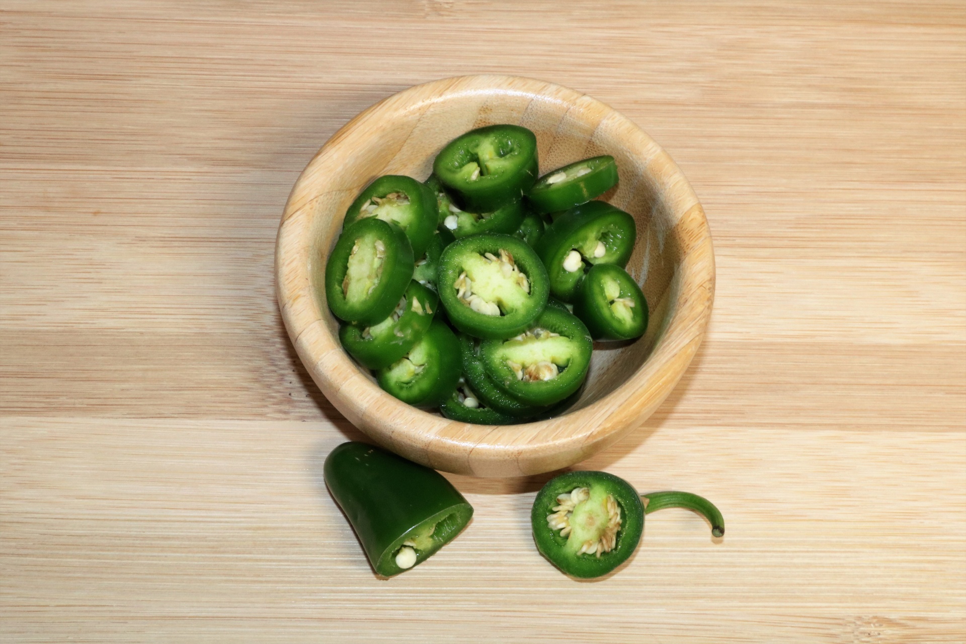 A small wooden bowl is filled with sliced green jalapeno peppers, sitting on a light brown wooden cutting board.