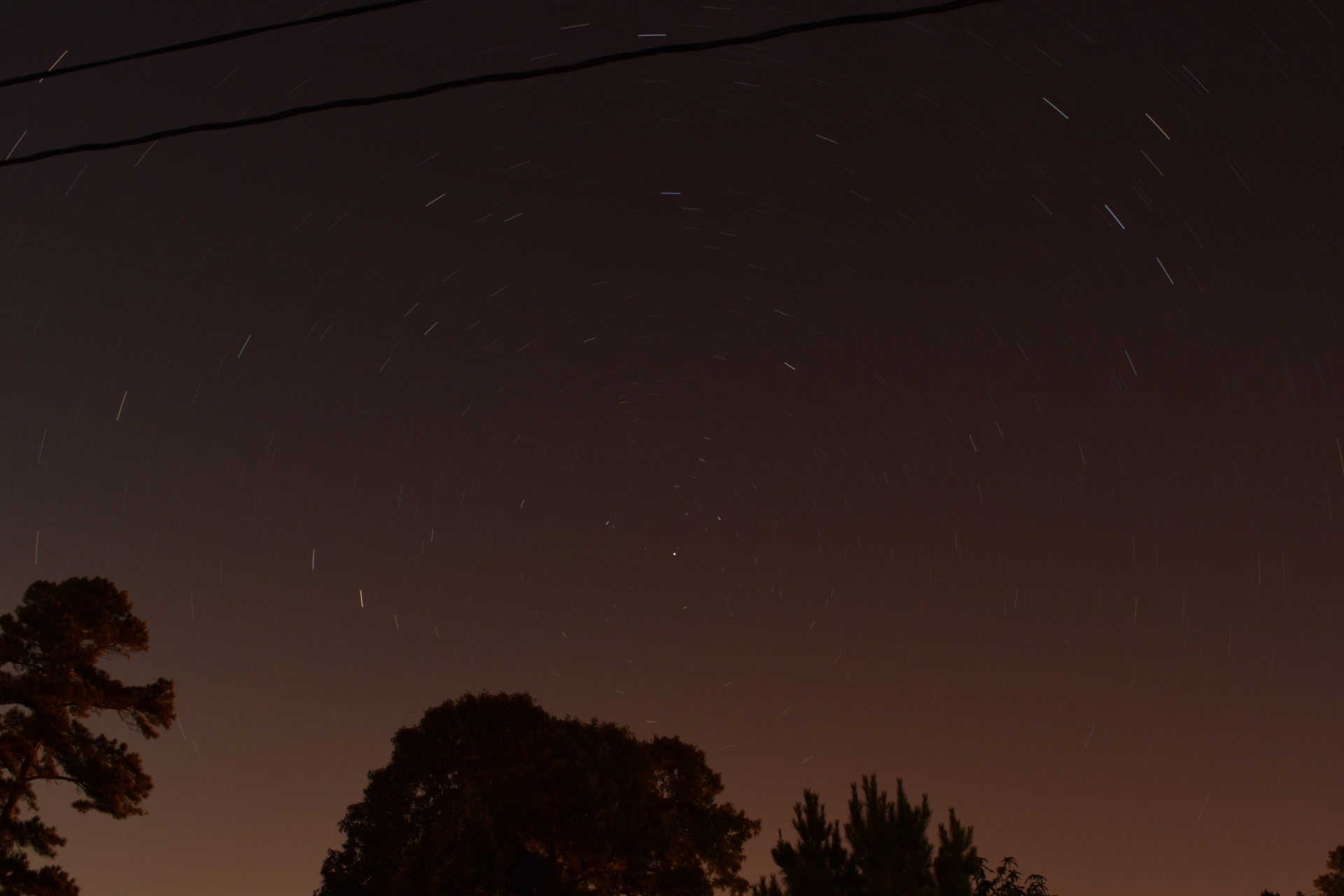 Stars forming a curving trail of light as the earth rotates, including some trees and a power line in the foreground. Polaris is not quite center, and is the star the others seem to spin around it. Single shot image, long exposure.