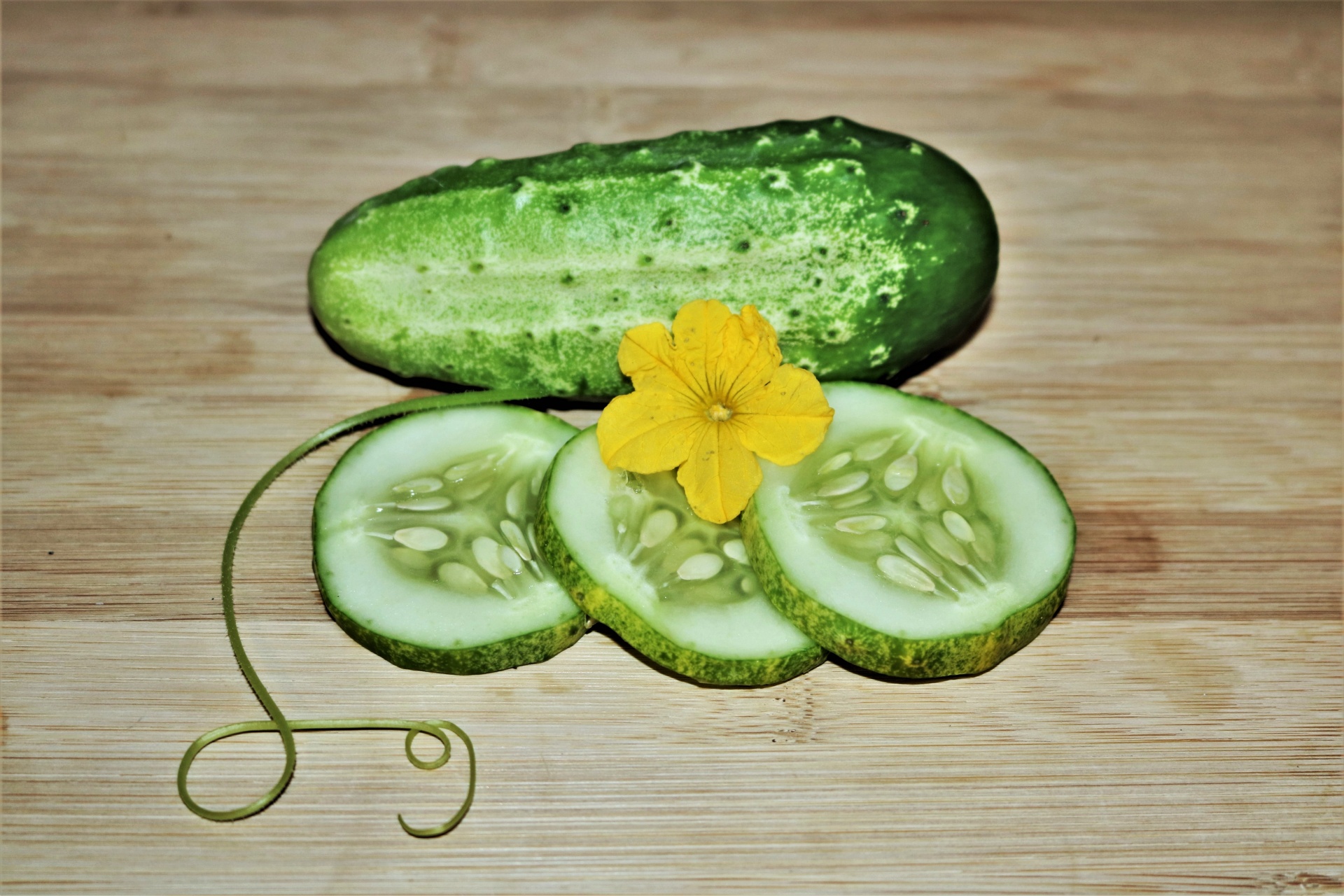 Cucumbers, sliced and whole, on a wood table, with cucumber bloom and tendril.