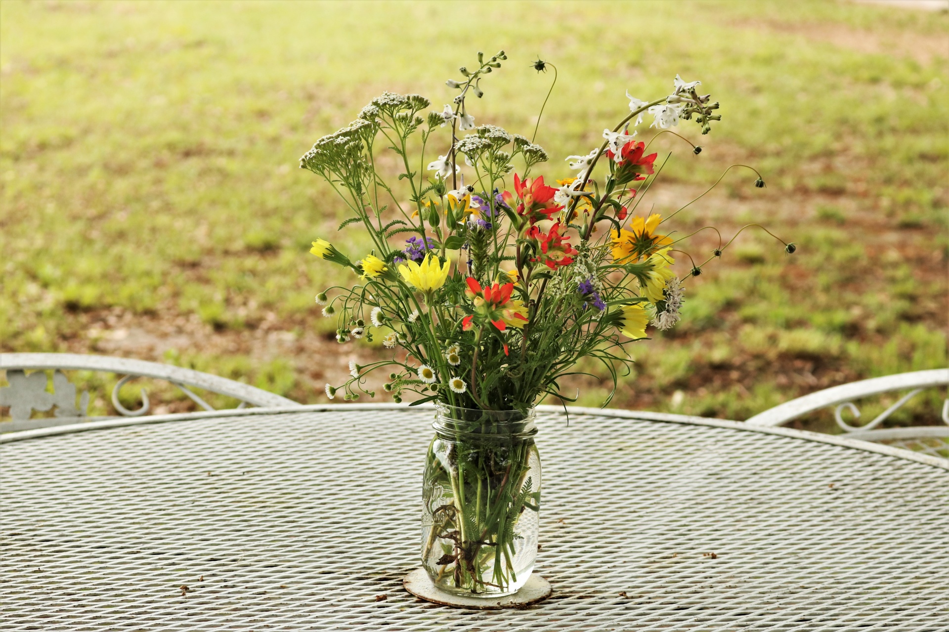 An assortment of wildflowers in a glass jar sitting outside on a white picnic table.
