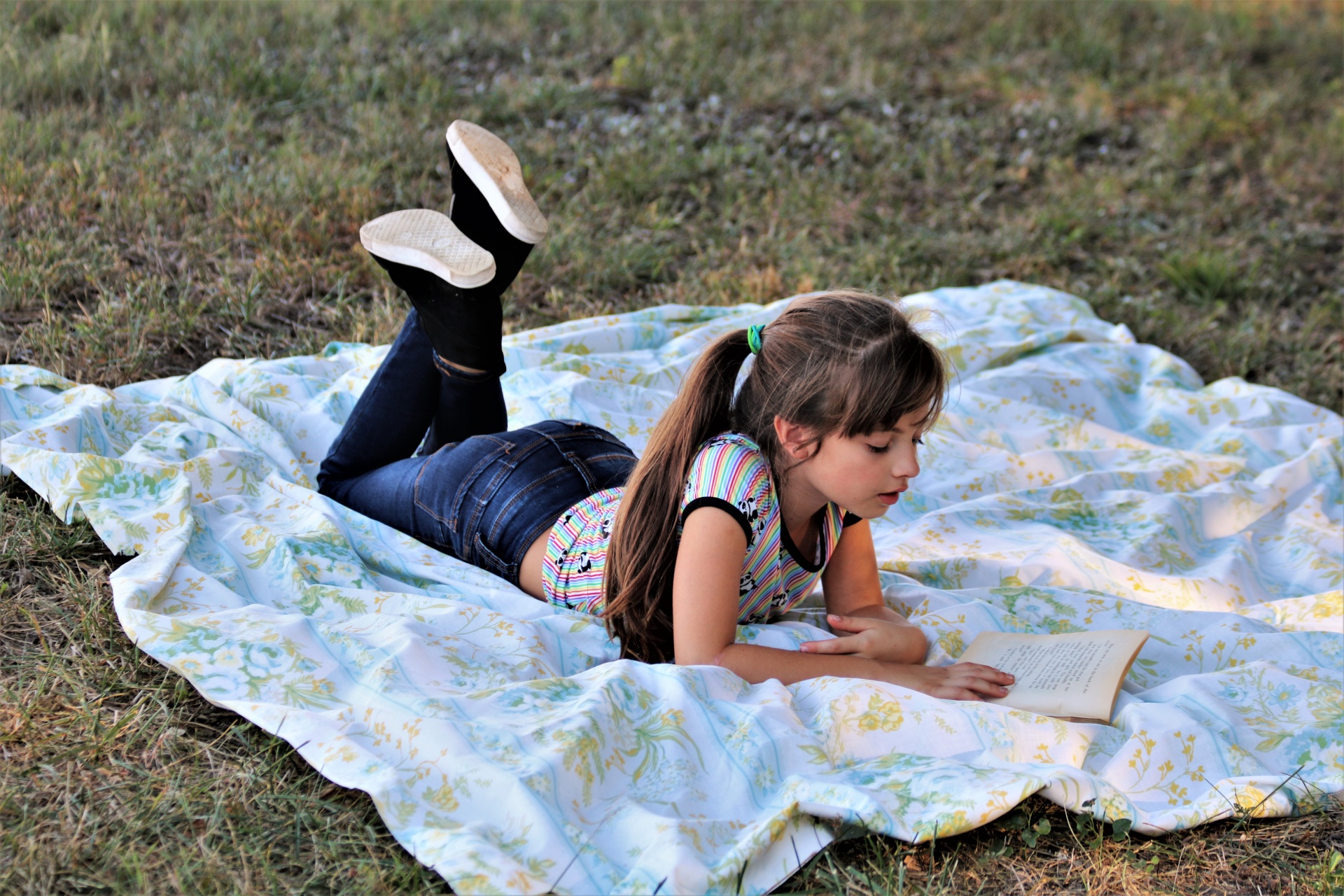 A young girl is lying on a blanket in the grass, reading a book.