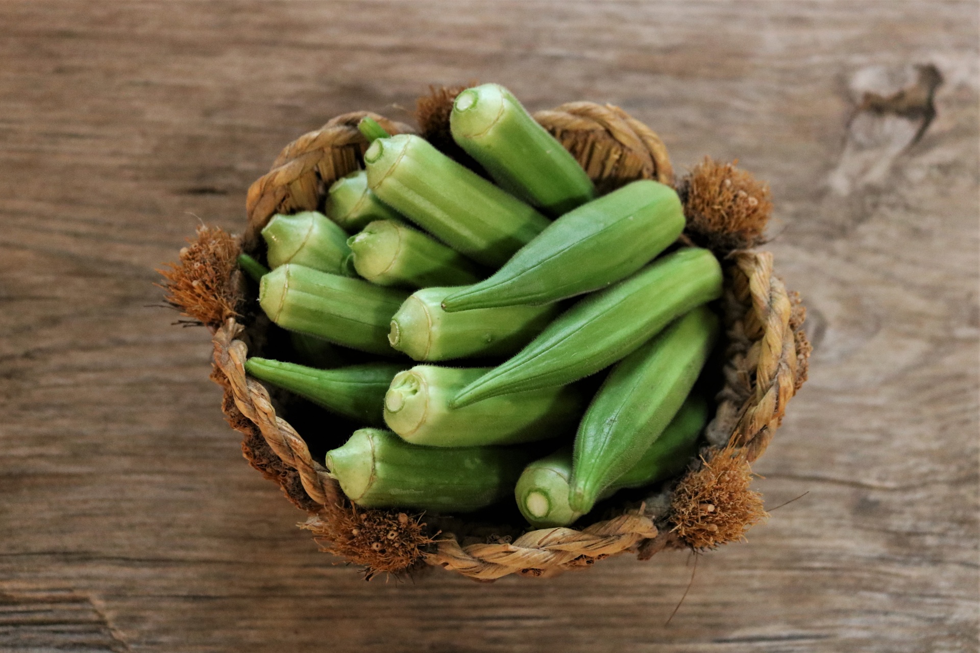 Top view of freshly picked okra in a straw basket on a wood table.