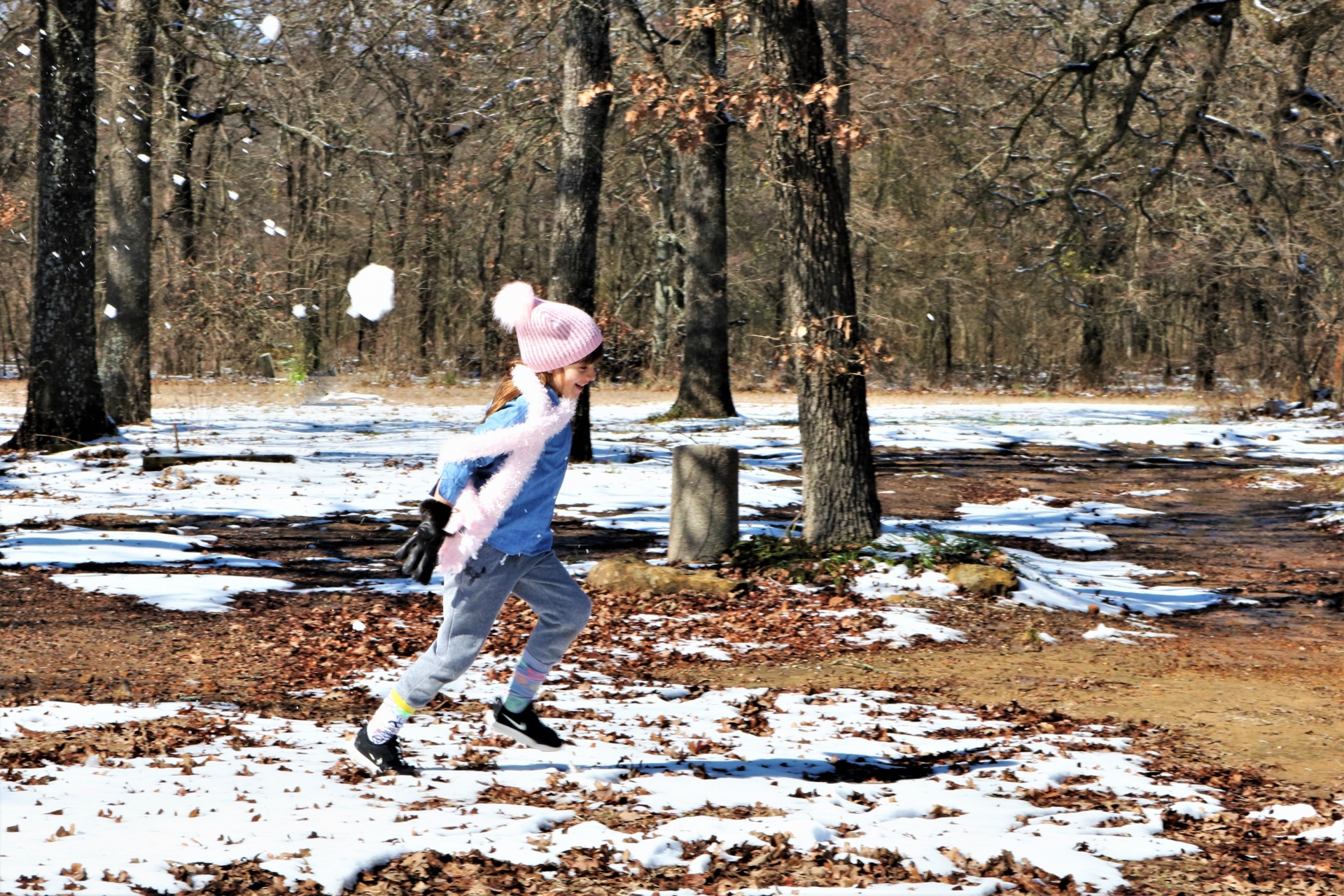 A cute little girl is running across the snow with a big snowball flying through the air behind her.