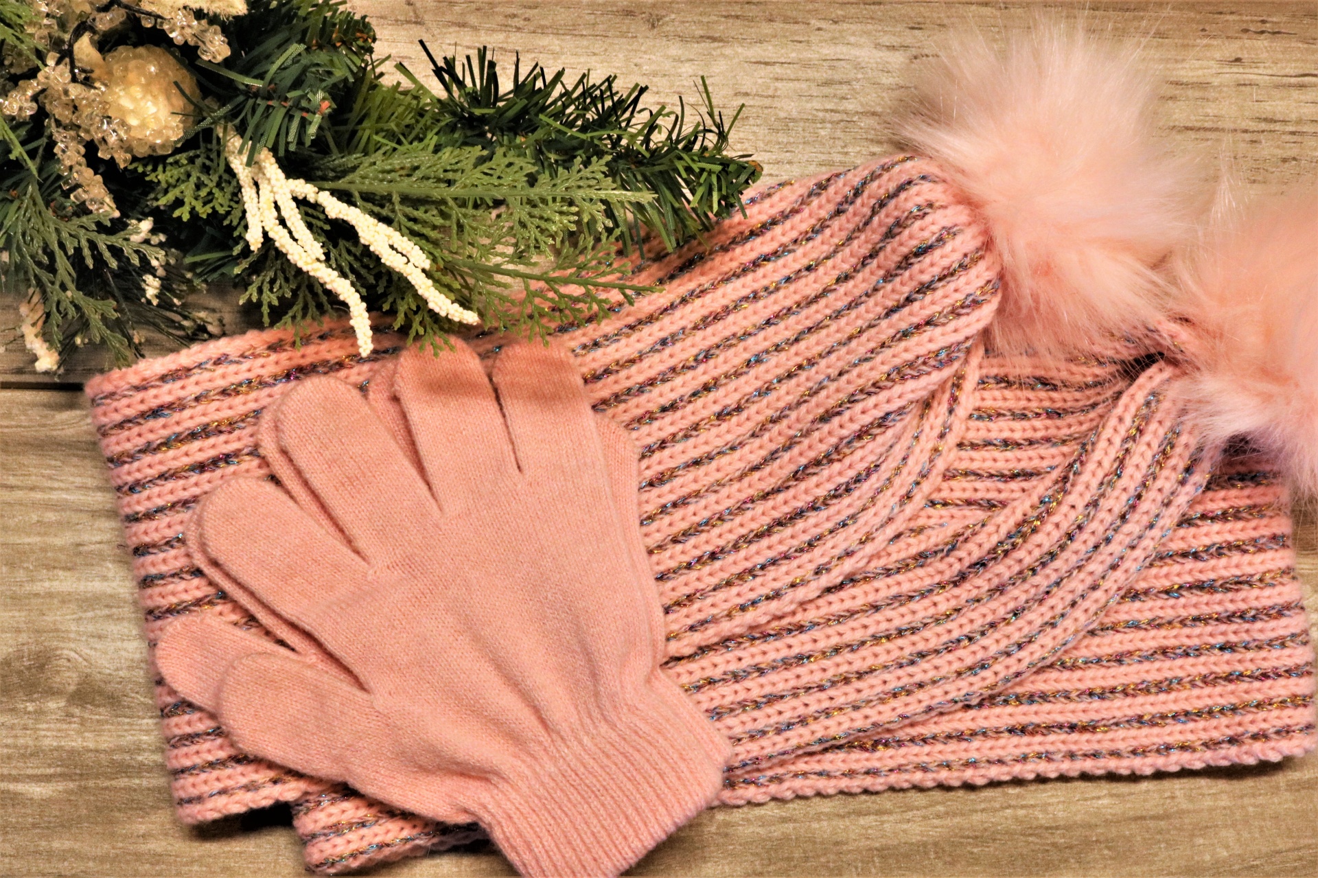 Close-up of a pink winter scarf, with silver stripes and a pair of pink gloves on a wood grain background with a Christmas tree branch in the background.