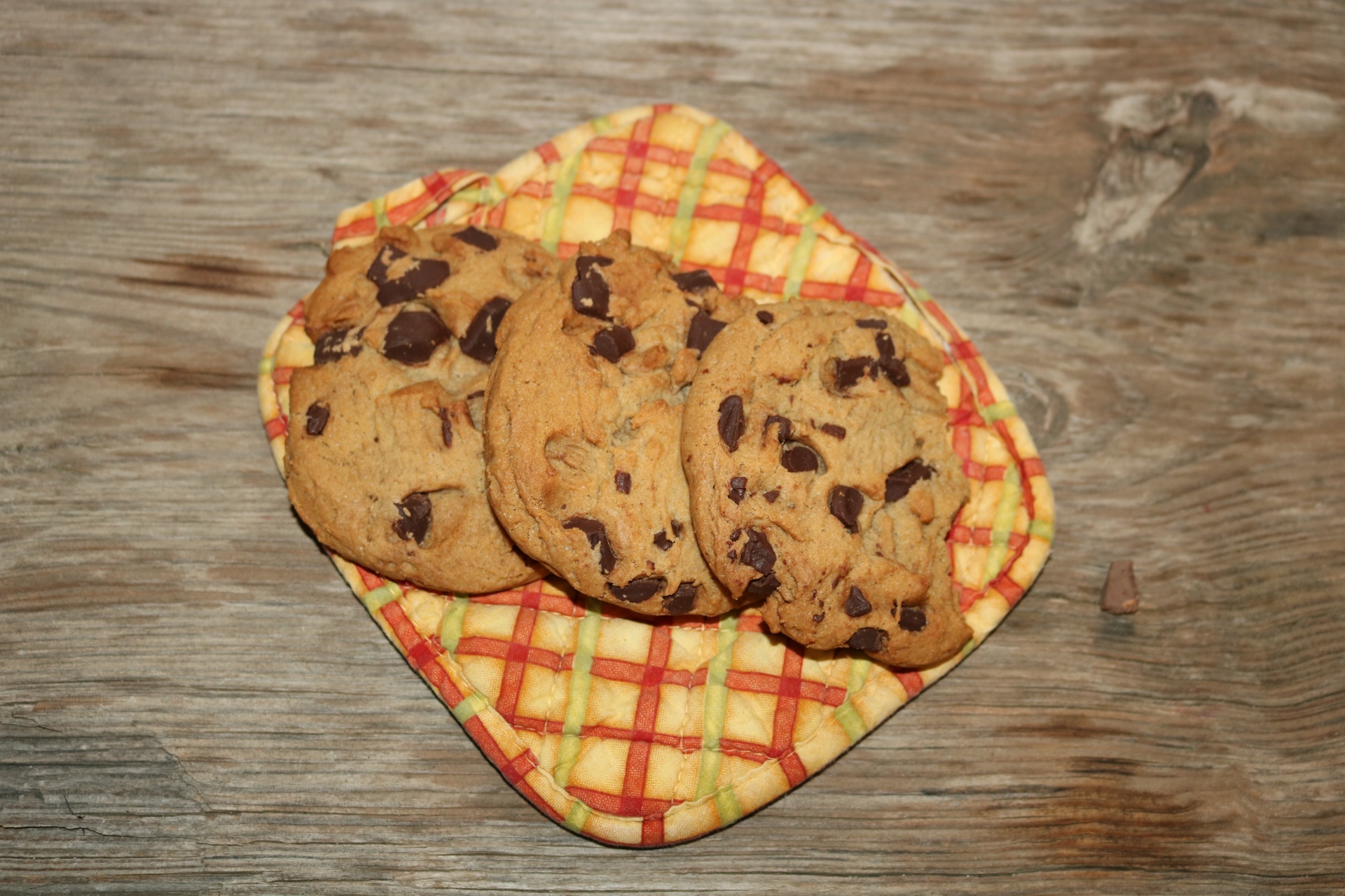 Top view of three chocolate chip cookies on a colorful pot holder, on a wood table.
