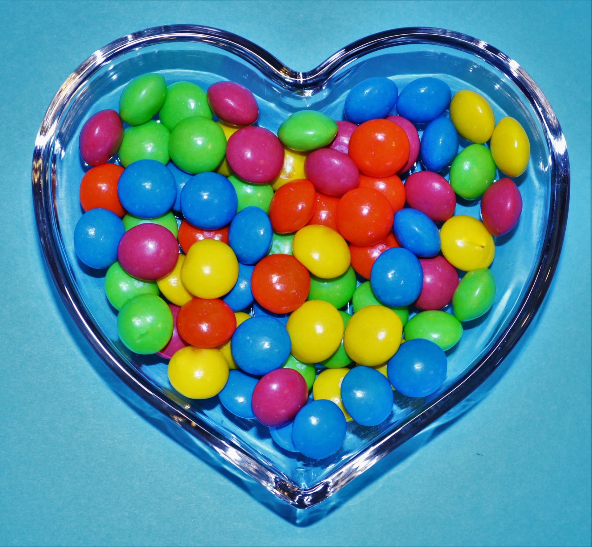 Top view close-up of colorful round candy in a heart dish, on a blue background.